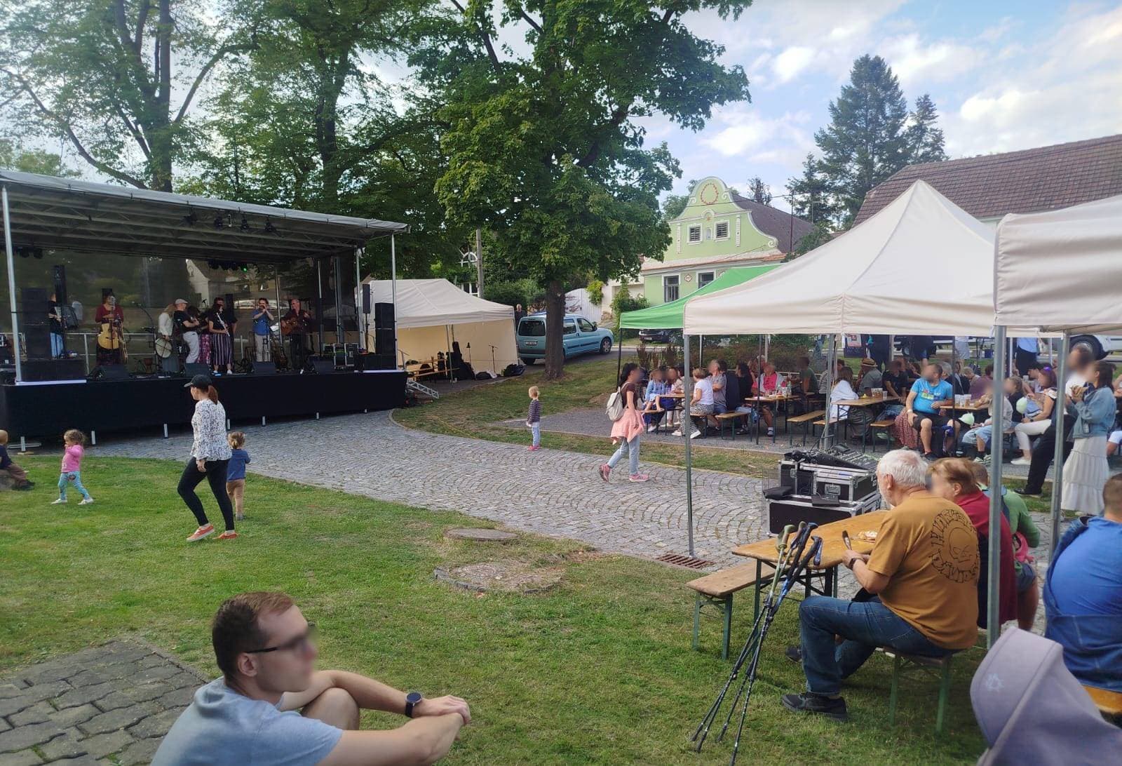 Akustická sestava na pódiu — bodhrán, píšťala a kytary. Před pódiem děti běhající po trávě